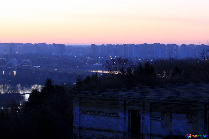 It shows an old building with a trees surrounding and some tall buildings in the background  Sunset on City №56010